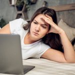 A girl lies on the bed and reads something on a laptop